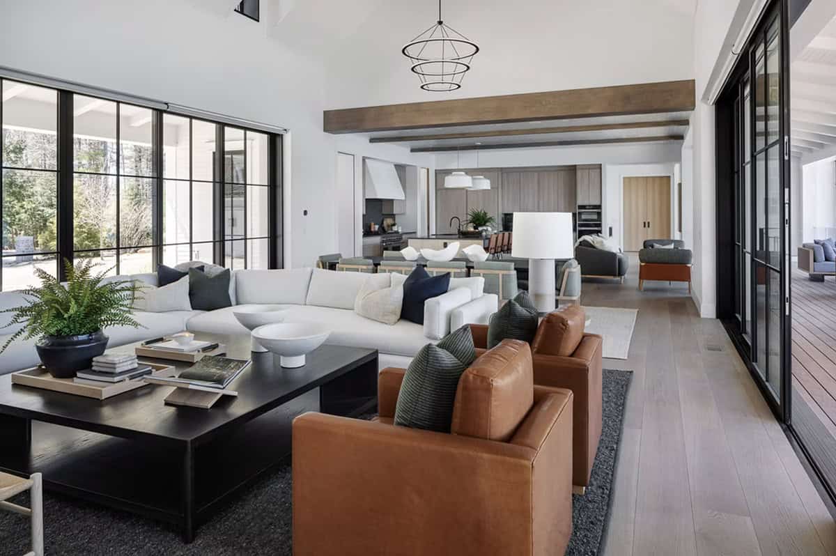 Expansive open-plan living area with cognac leather armchairs, white sectional, exposed wood beams, and lake-facing glass walls