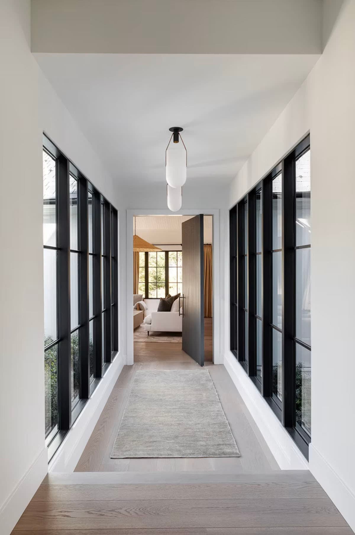 Glass-lined interior corridor with black-framed windows on both sides, white oak floors, and frosted pendant light