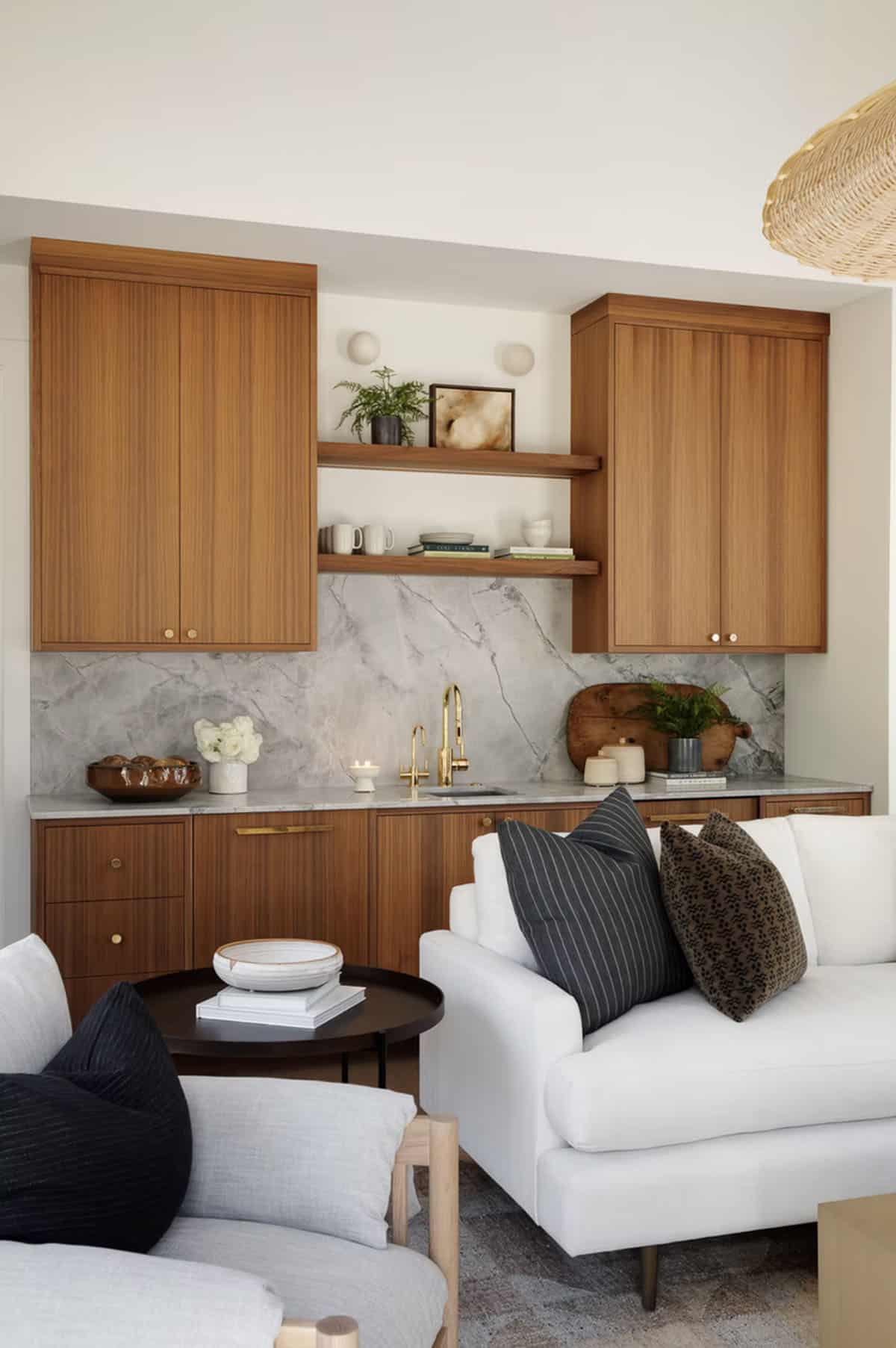 Warm-toned wet bar with walnut cabinetry, marble backsplash, brass faucet, open shelving, and white sofa in foreground