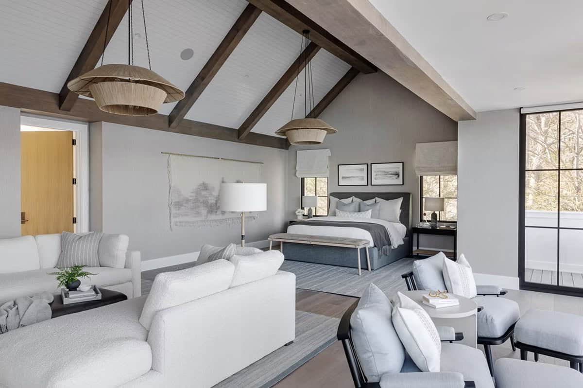 Spacious primary suite with cathedral ceiling, exposed wood beams, dual rattan pendants, gray upholstered bed, and sitting area