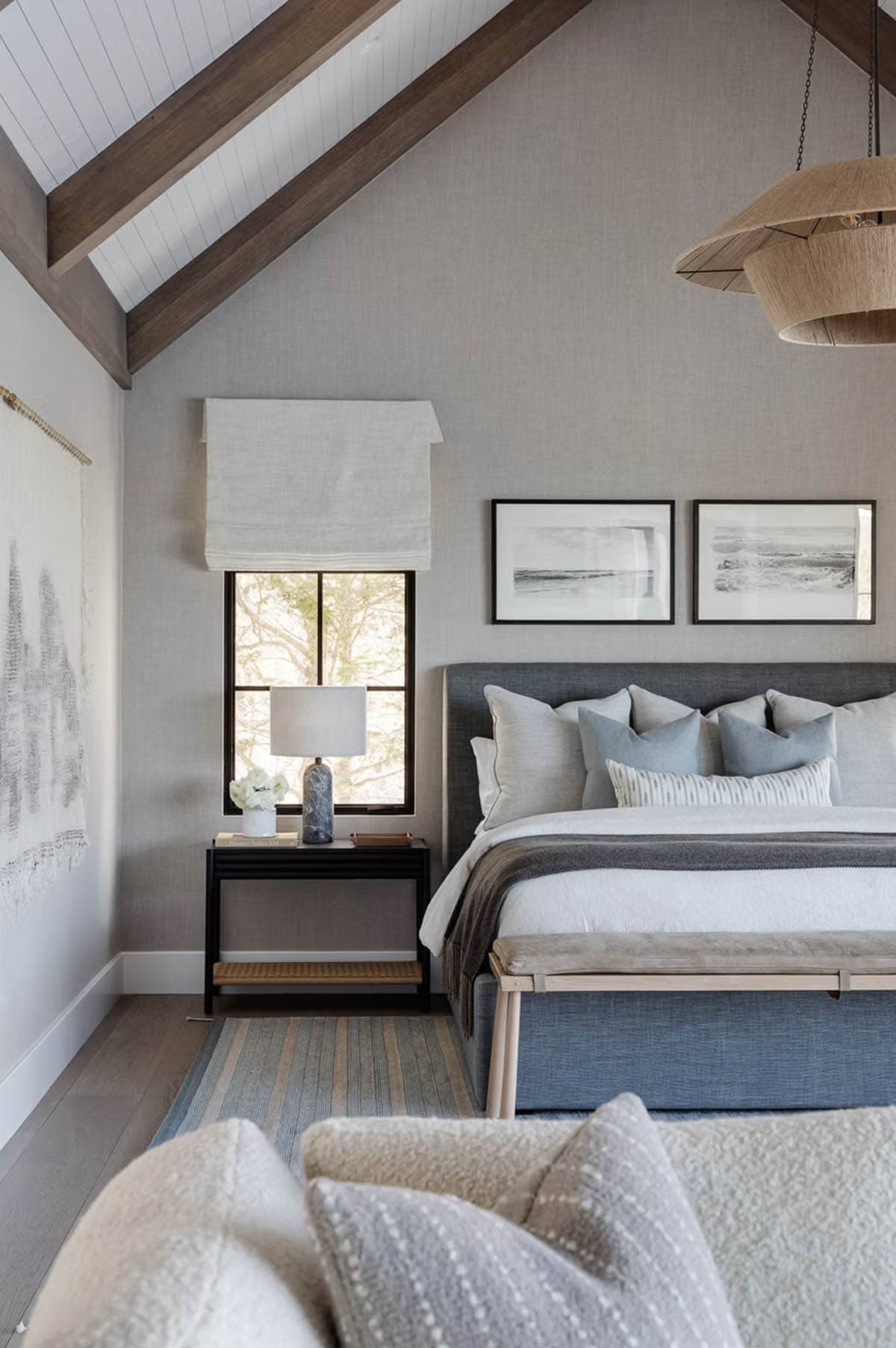 Primary bedroom detail with vaulted shiplap ceiling, gray linen upholstered bed, black-and-white landscape art, and rattan pendant