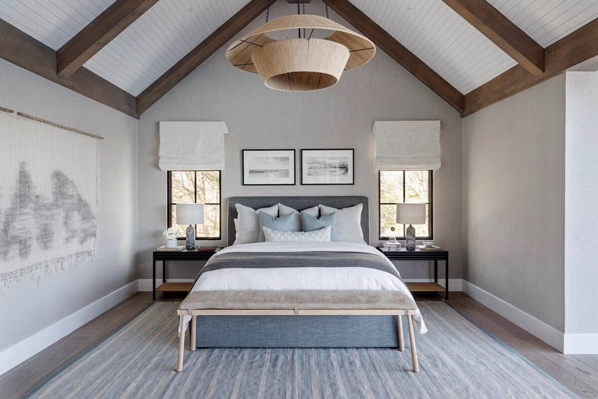 Primary bedroom with cathedral ceiling, exposed beams, stacked rattan chandelier, gray upholstered bed flanked by black nightstands