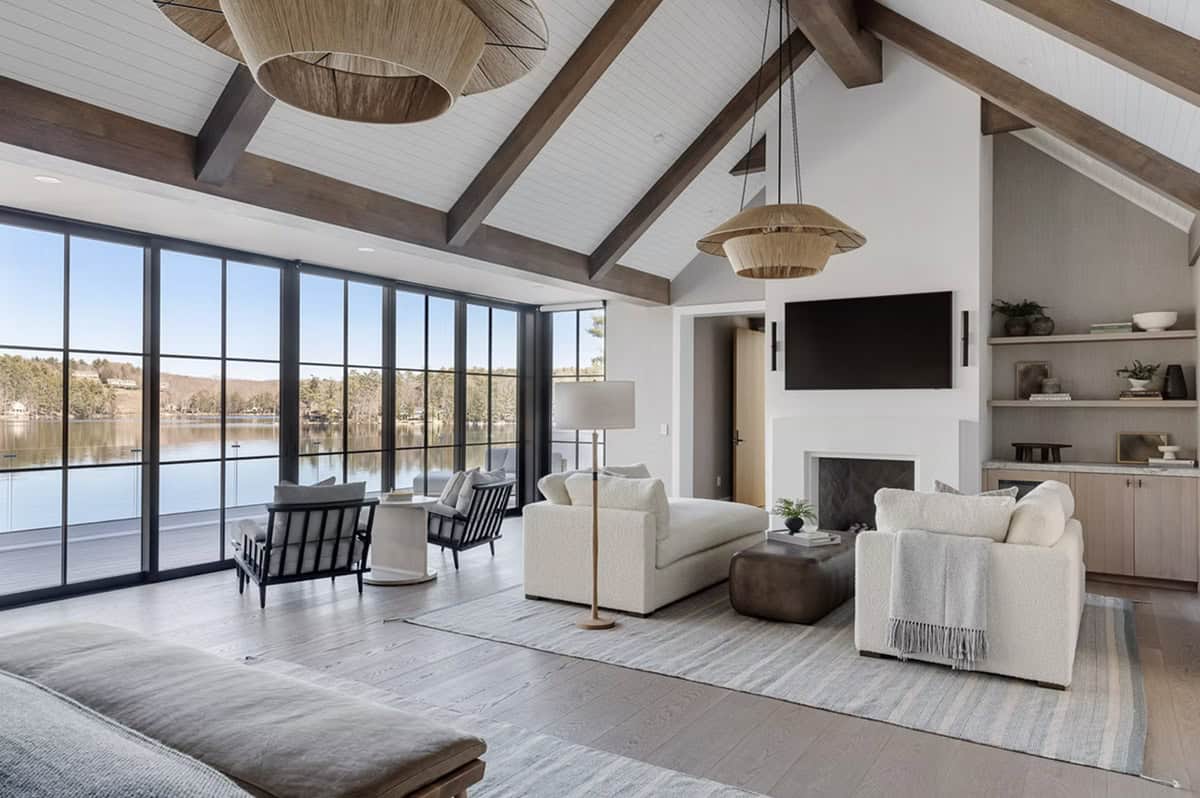 Vaulted primary suite sitting area with rattan pendants, floor-to-ceiling lake views, fireplace, built-in shelving, and TV