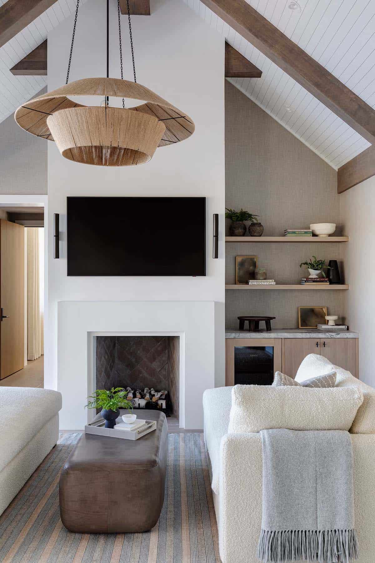 Primary suite fireplace wall with plaster surround, TV, floating oak shelves, leather ottoman, and large rattan chandelier