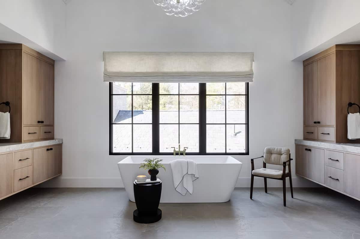 Spa-like primary bathroom with freestanding soaking tub, dual floating oak vanities, marble countertops, and black-framed window