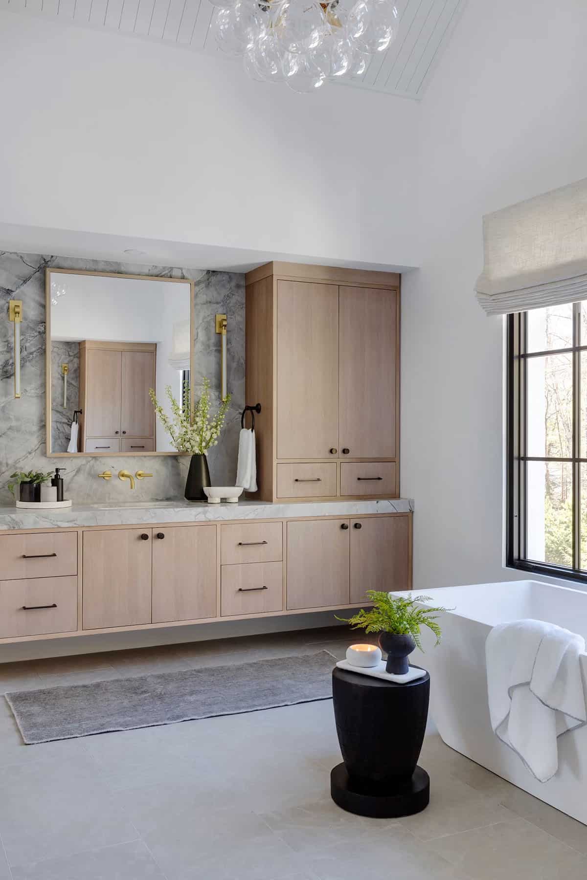 Primary bathroom with floating oak vanity, marble slab backsplash, gold sconces and faucets, and bubble glass chandelier