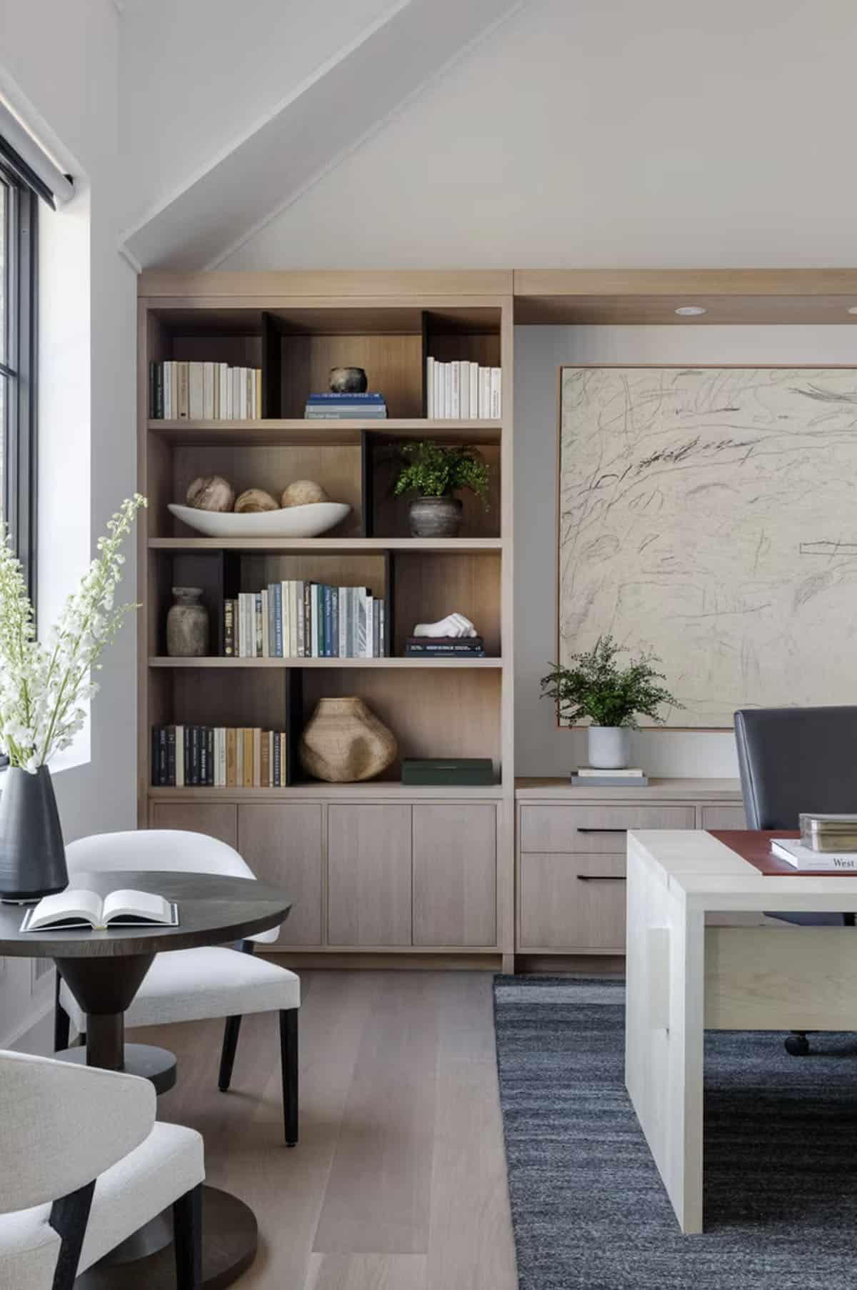 Home office with styled light oak built-in shelving, large abstract artwork, round side table, and white accent chairs