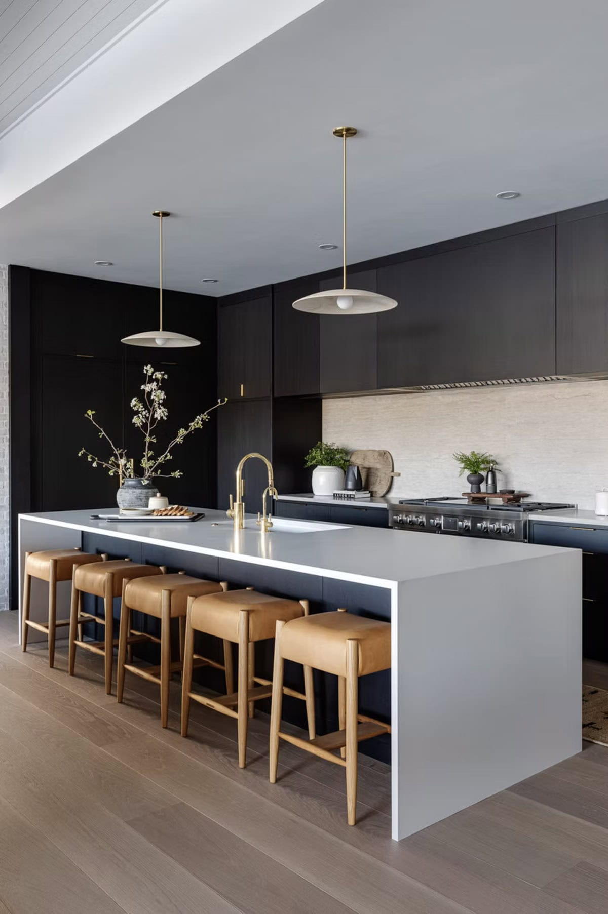 Sleek lower-level kitchen with matte black cabinetry, white waterfall island, brass fittings, leather counter stools, and saucer pendants