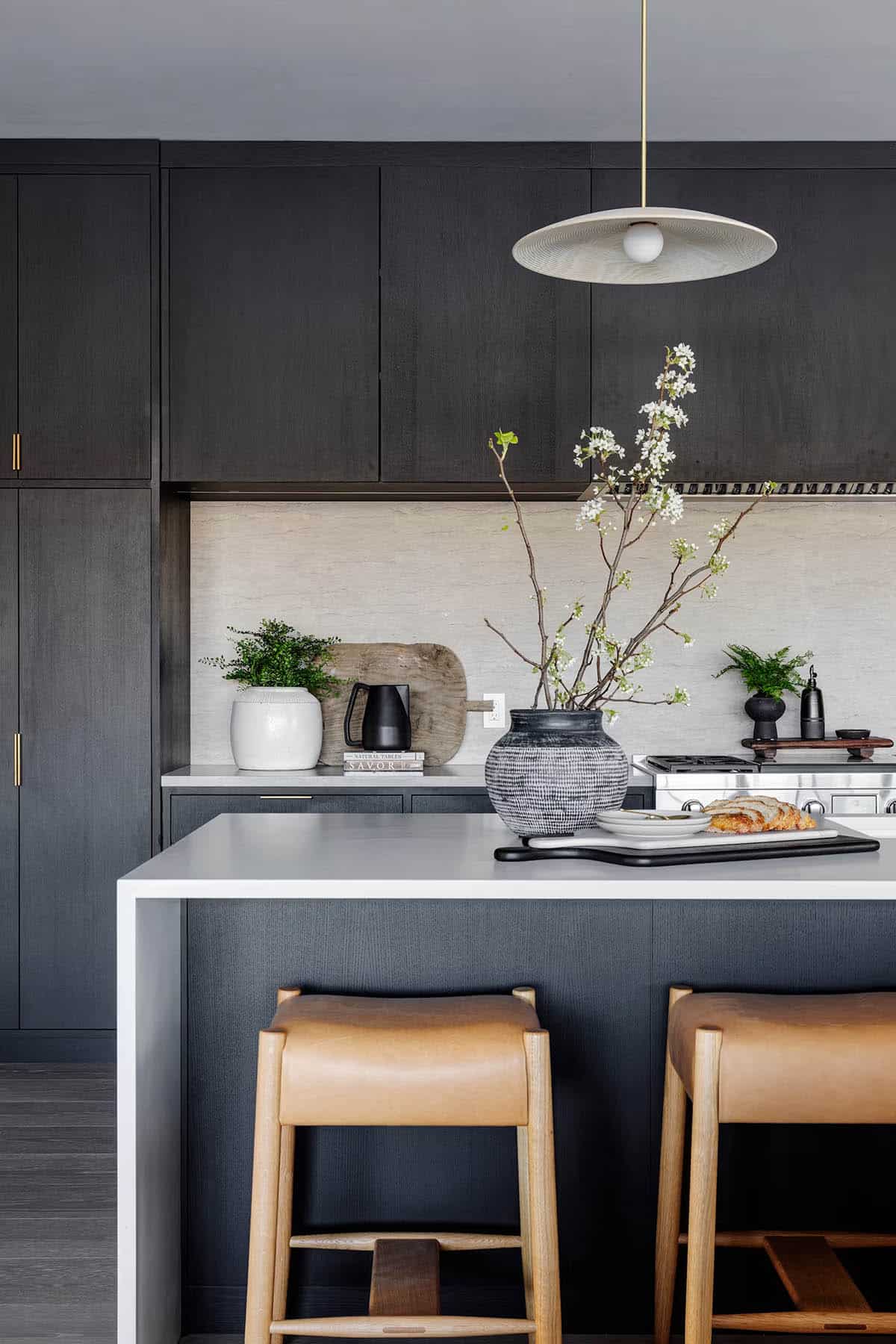 Lower-level kitchen island detail with charcoal cabinetry, white quartz countertop, leather bar stools, stone backsplash, and saucer pendant