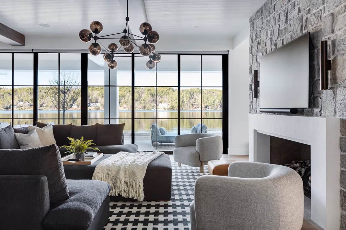Lower-level living room with dark sectional, stone fireplace, TV, globe chandelier, and panoramic lake views through steel-framed glass walls