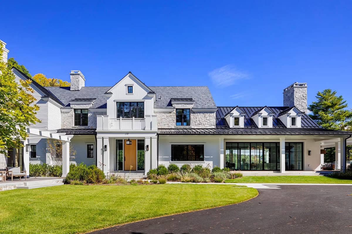 Street-facing facade of contemporary lakeside home with natural wood front door, stone and white exterior, slate and metal roof, circular driveway