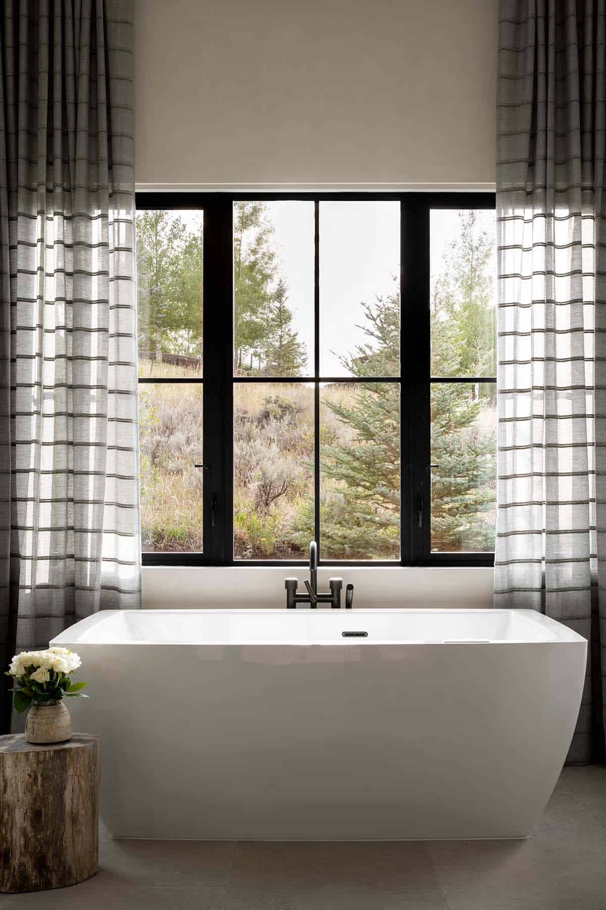 Freestanding soaking tub beneath black-frame window with sage and pine tree views