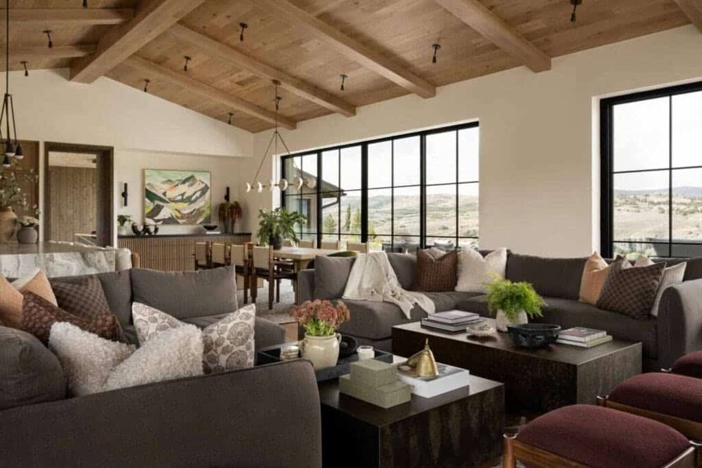 Open living room with gray sectionals, wood beam ceiling, and black-frame mountain views