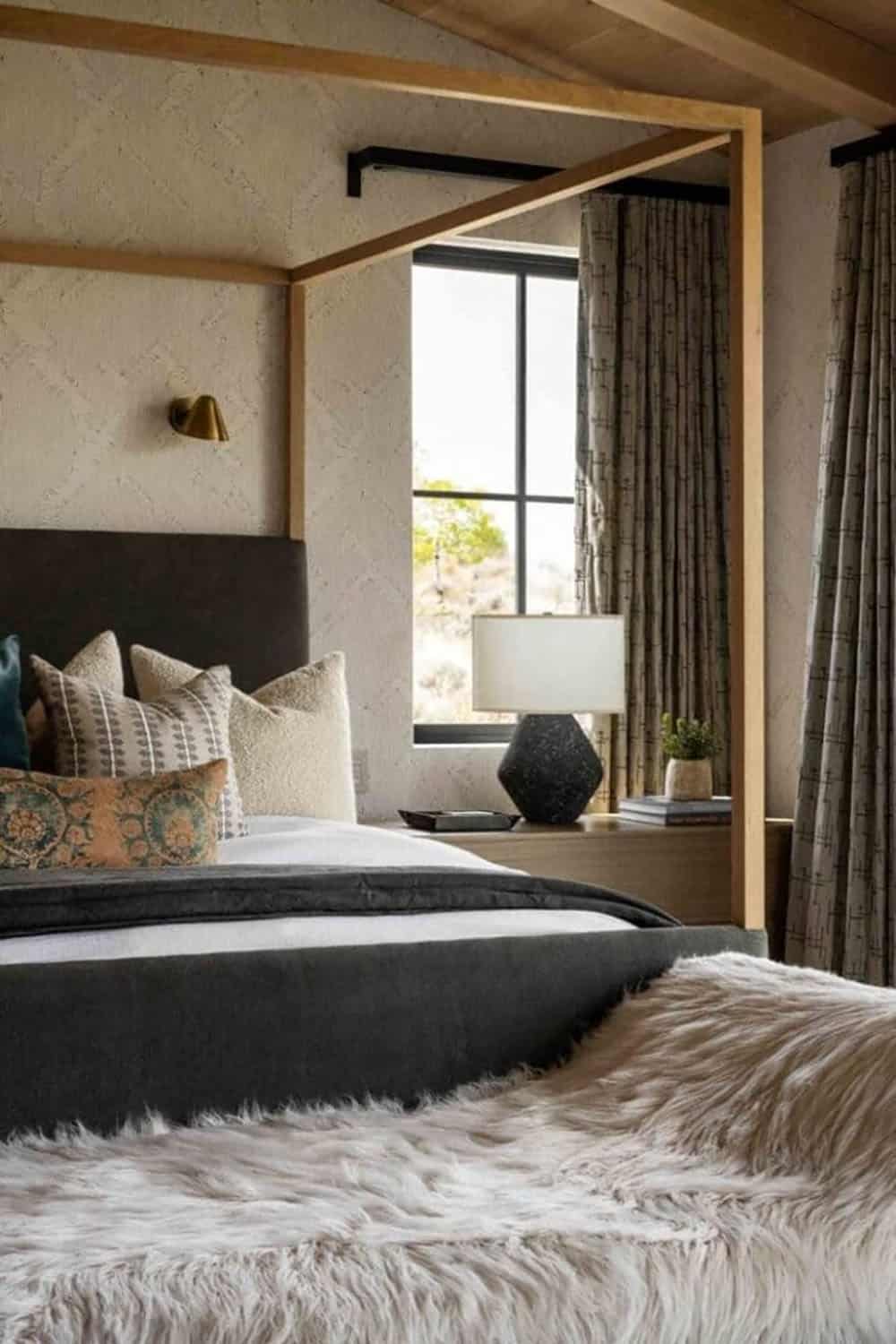 Primary bedroom close-up with wood canopy bed, layered pillows, fur throw, and brass sconce