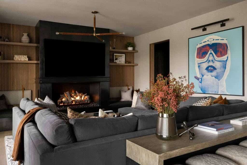 Lower-level lounge with dark sectional sofas, steel fireplace, ski pop-art painting, and brass chandelier