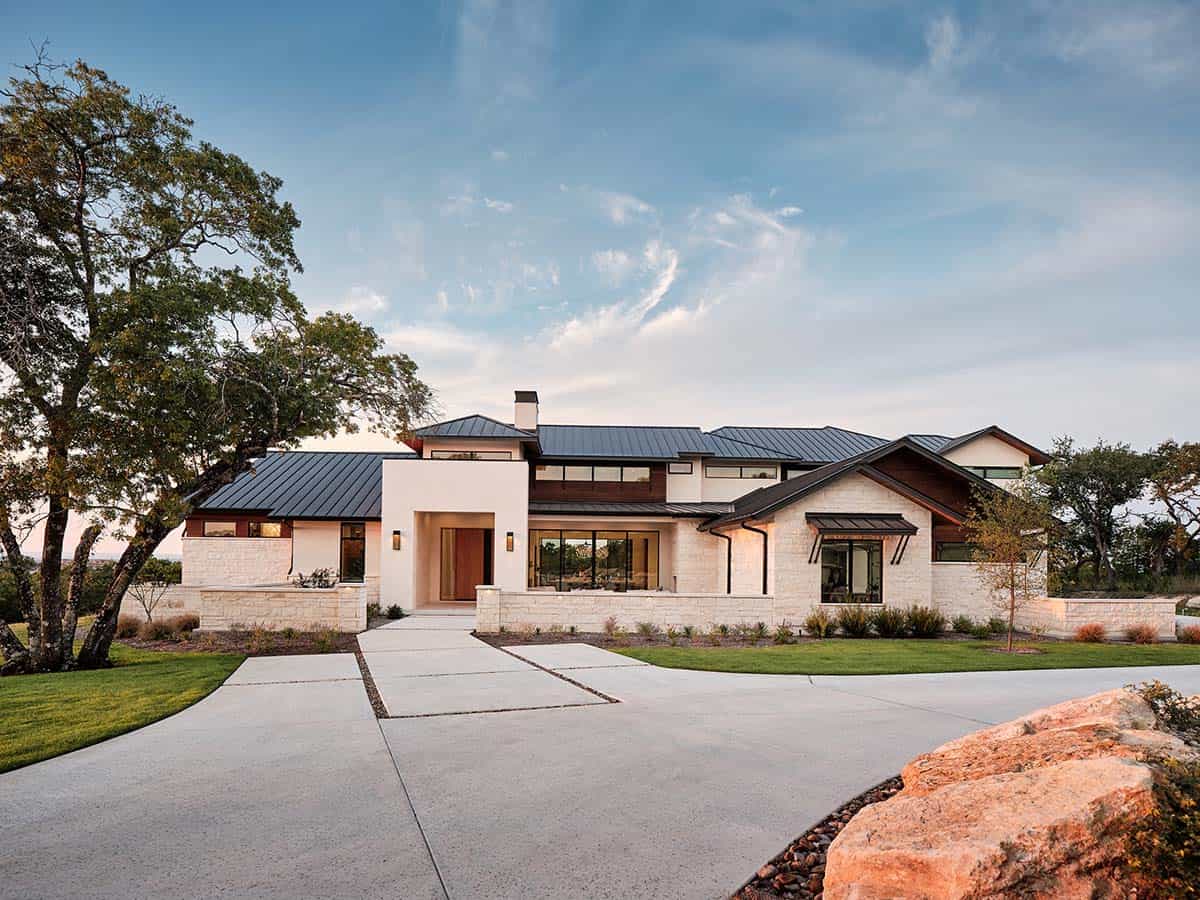 Luxury organic modern home exterior with limestone, white stucco, dark metal roof, and oak trees in Texas