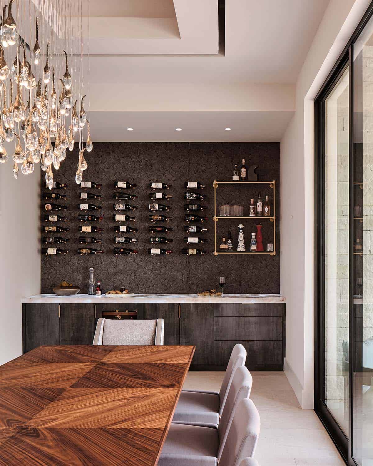 Dining room wine wall with dark textured tile, wall-mounted bottle racks, brass bar shelving, and crystal chandelier