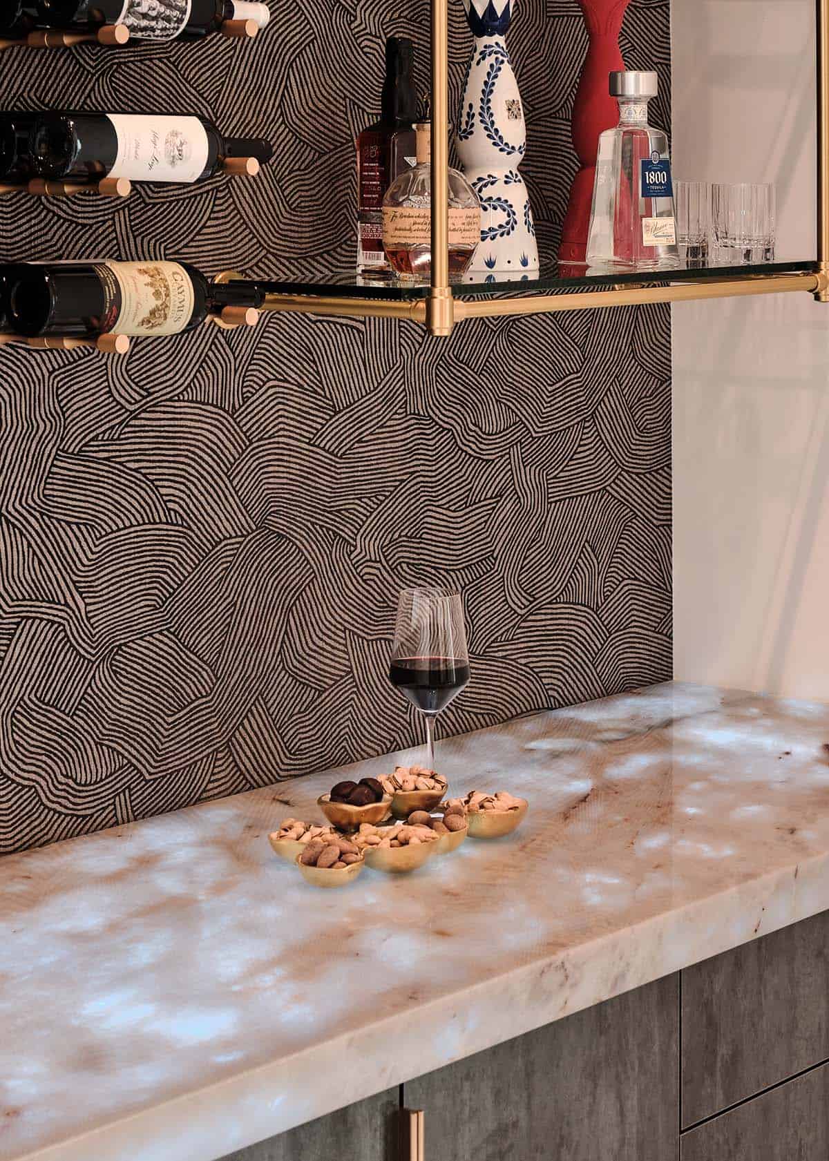 Bar countertop detail with onyx surface, wine bottles, brass shelving, and dark patterned tile backsplash