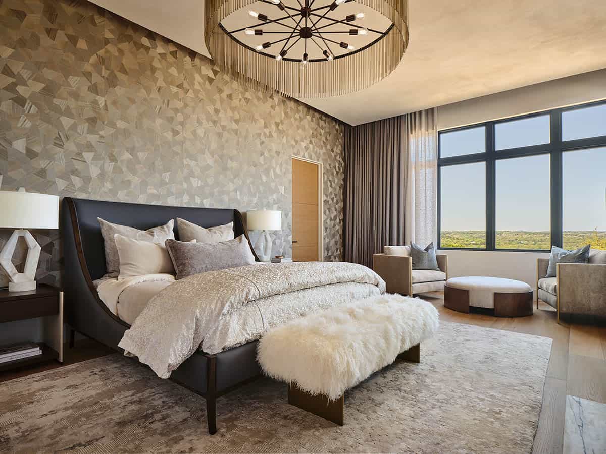 Luxury primary bedroom with geometric wallpaper accent wall, dark leather bed, fringe chandelier, and hill country views