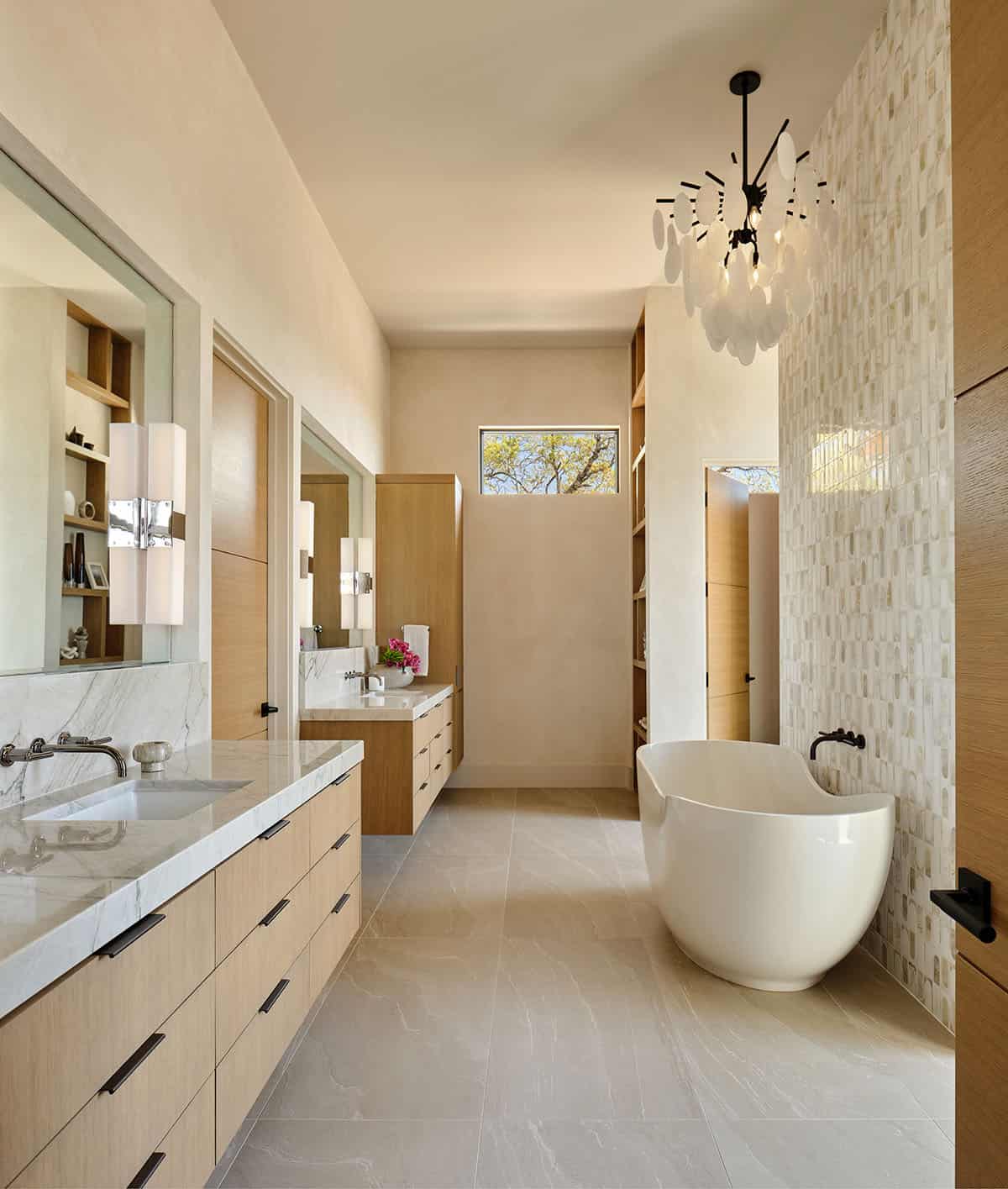 Primary bathroom with oak floating vanities, freestanding soaking tub, mosaic tile accent wall, and sculptural chandelier