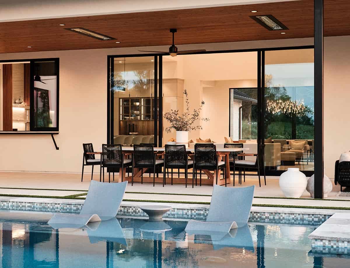 Pool with in-water lounge chairs and covered outdoor dining area viewed through floor-to-ceiling glass doors at dusk