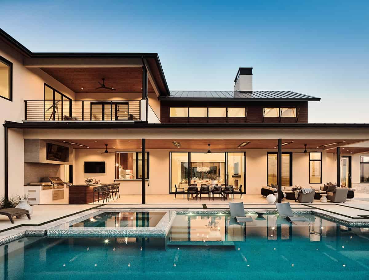Luxury home rear exterior at dusk with infinity pool, spa, covered patio, outdoor kitchen, and upper balcony
