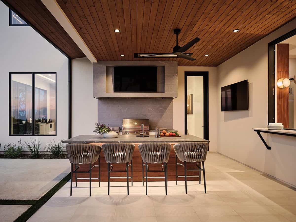 Covered outdoor kitchen with stone island, grill, woven barstools, wood plank ceiling, and outdoor TV