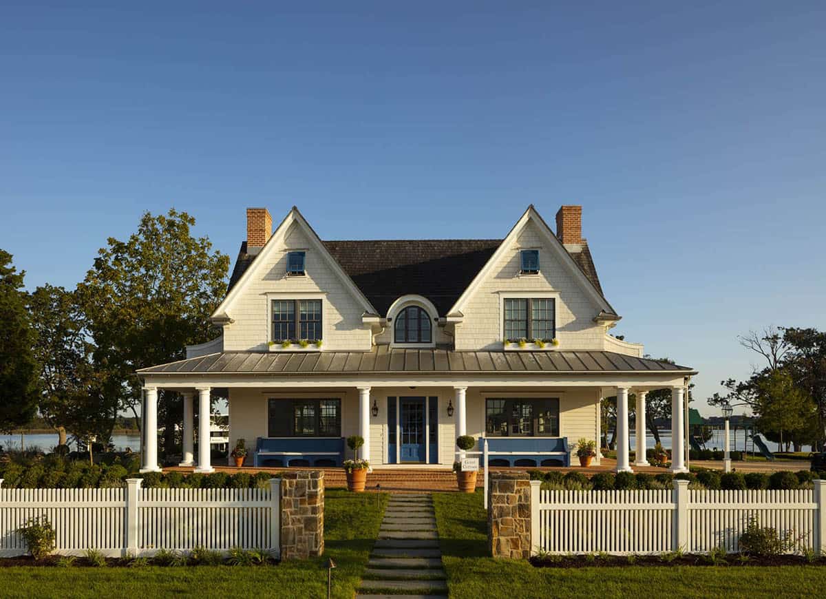 Front facade of white shingle-style guest cottage at golden hour with covered porch, blue front door, twin brick chimneys, and picket fence