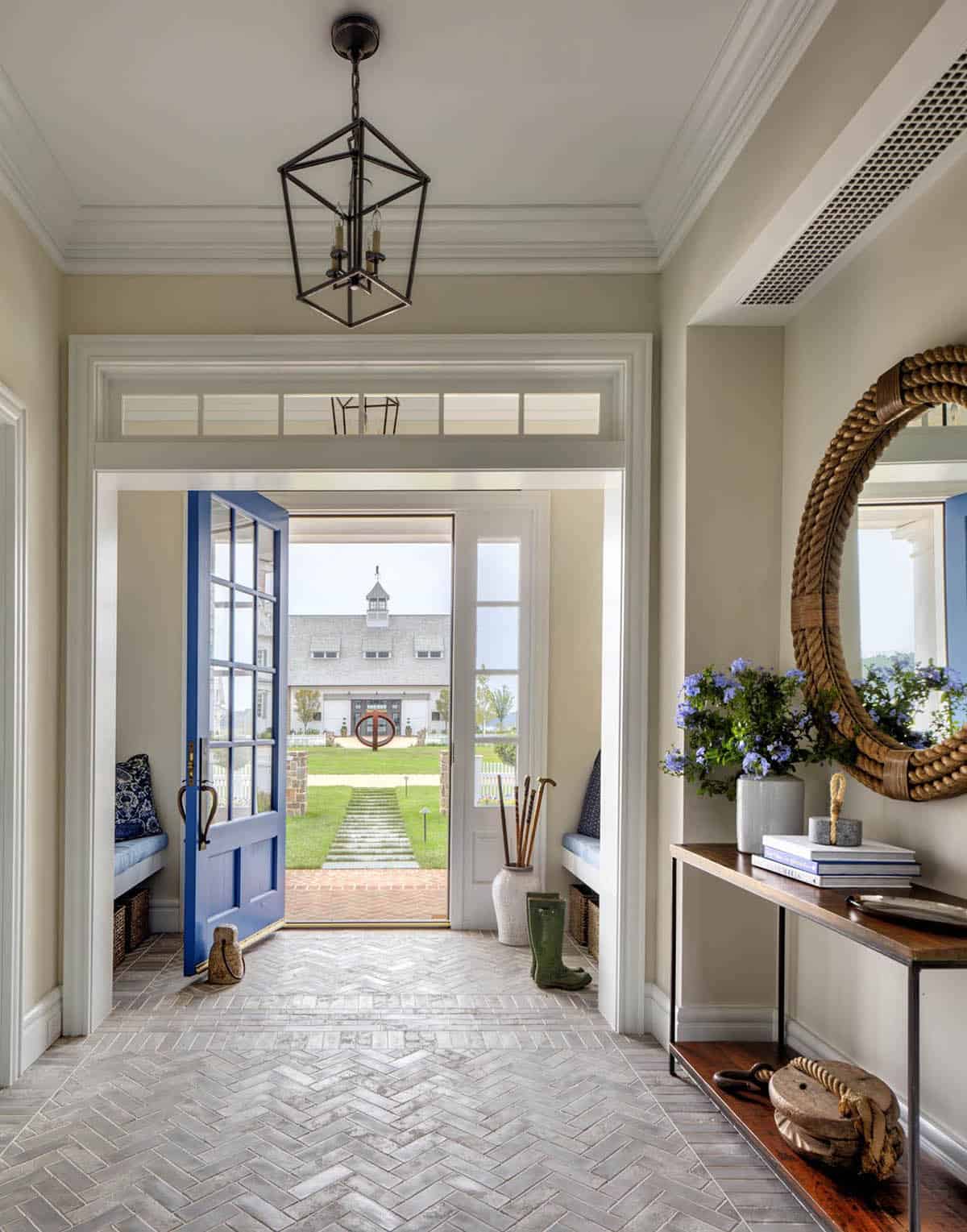 Guest cottage entry foyer with herringbone tile floor, open blue French door, iron lantern pendant, rope-frame mirror, and console table