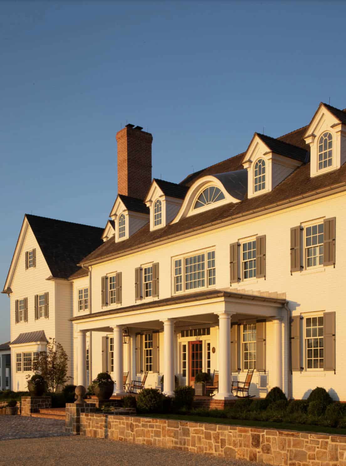 Close-up of manor house facade at sunset showing columned front porch, dormer windows, brick chimneys, and gray shutters