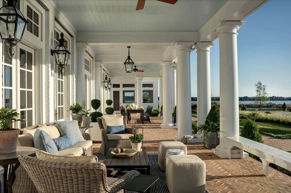 Covered rear porch with white Ionic columns, wicker seating, herringbone brick floor, and panoramic waterfront views
