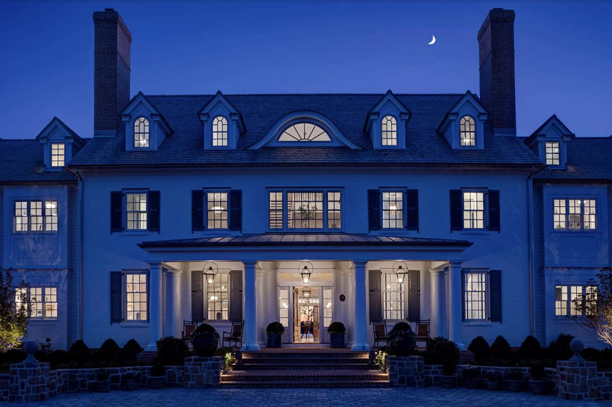 Front facade at twilight with lit windows, gas lanterns glowing under portico, crescent moon above twin brick chimneys