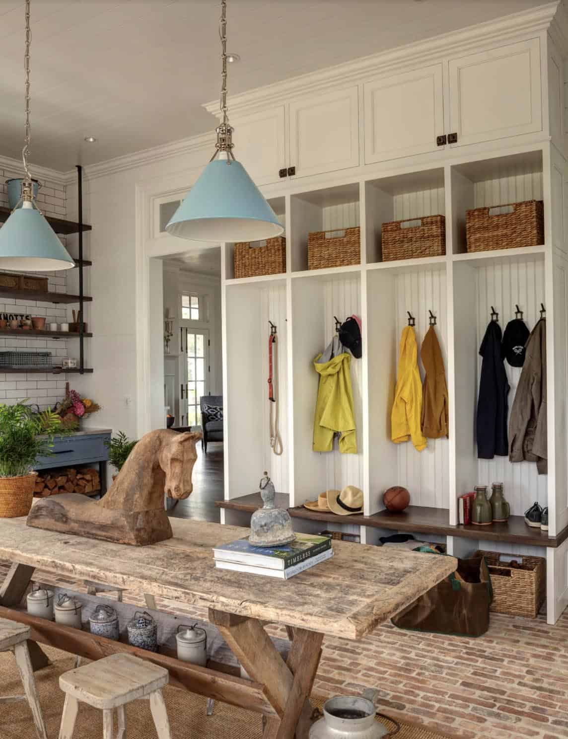 Mudroom with built-in white locker storage, wicker baskets, rustic worktable, carved horse sculpture, and blue pendant lights