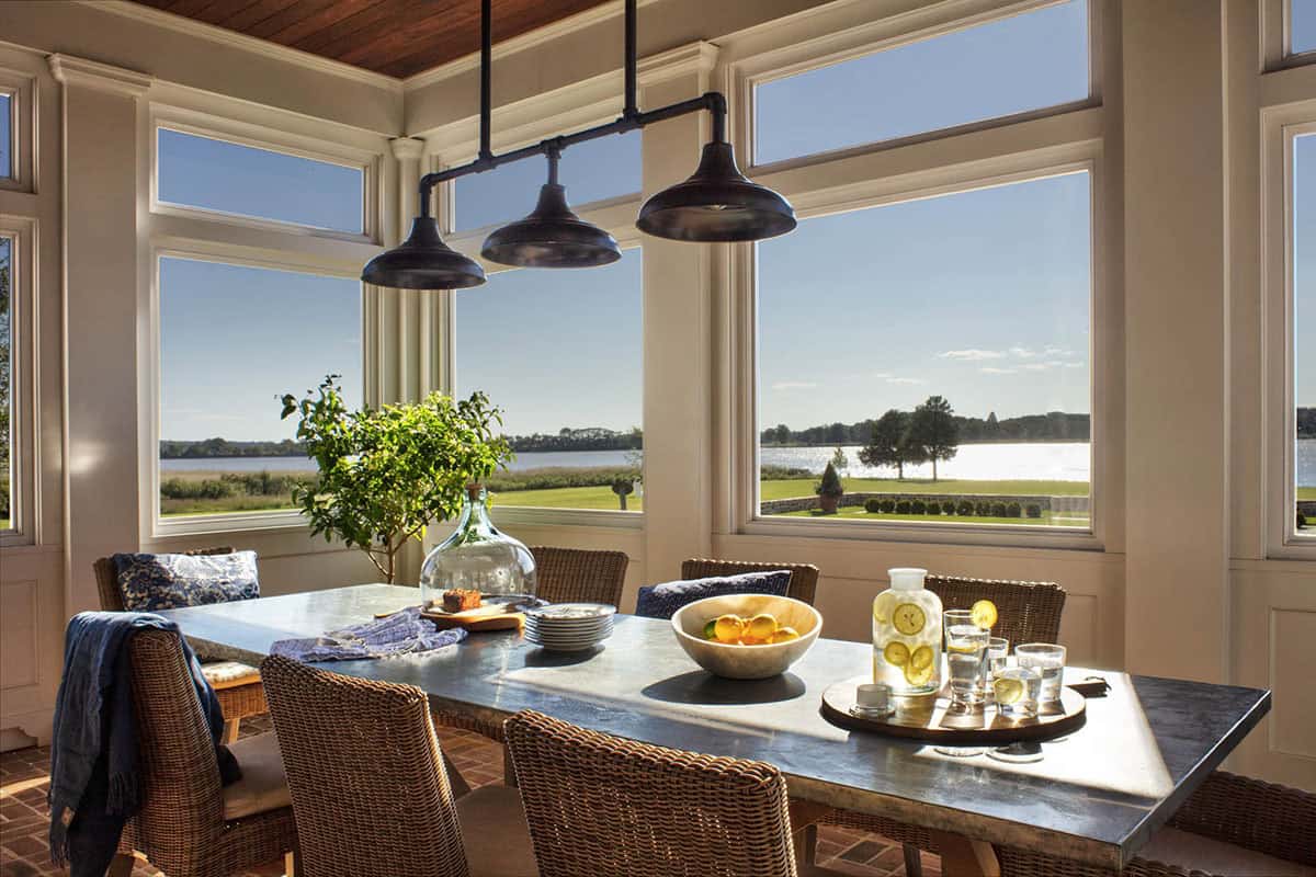 Sunlit breakfast nook with wicker chairs, zinc-top table, triple pendant lights, and expansive windows overlooking the river