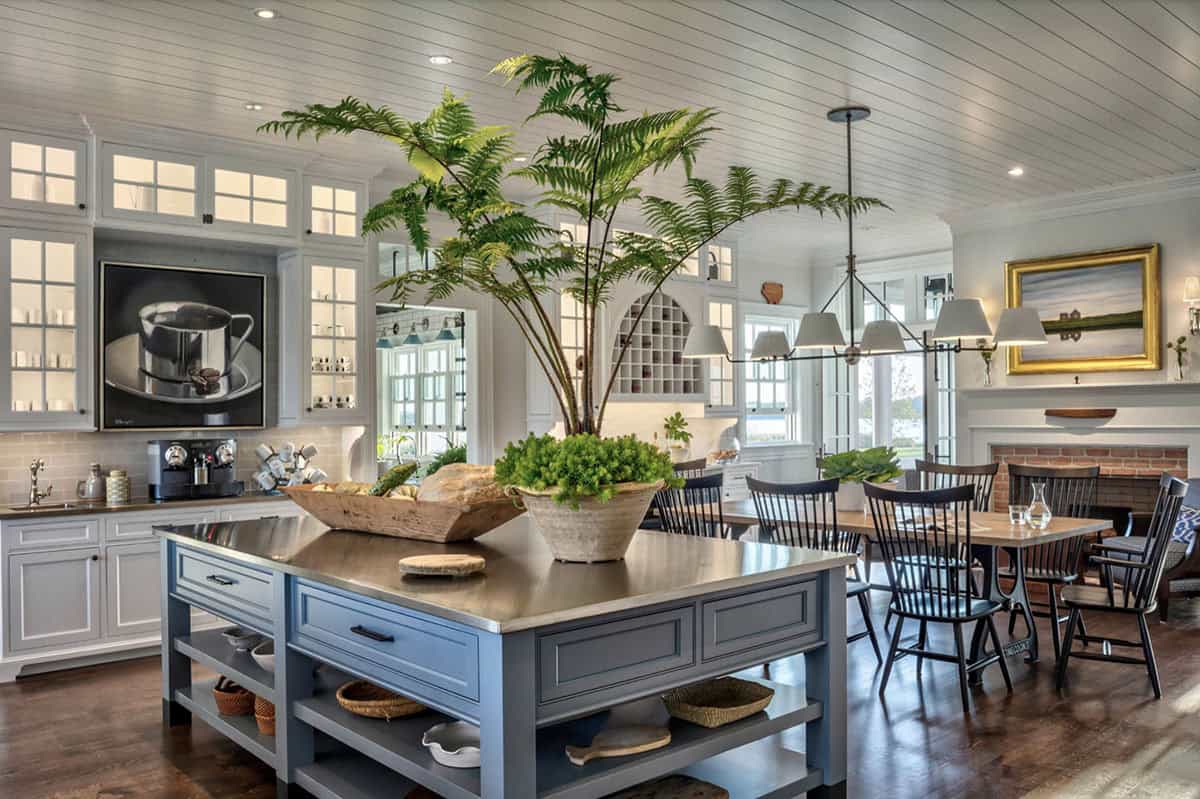 Open-plan kitchen with blue-gray island, large tree fern centerpiece, white cabinetry, and adjoining dining area with brick fireplace
