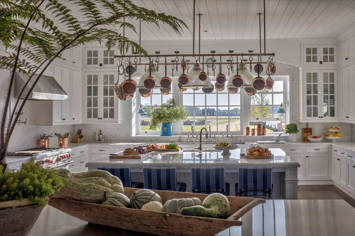 Grand kitchen with white cabinetry, hanging copper pots, large picture window overlooking the estate, and blue island stools