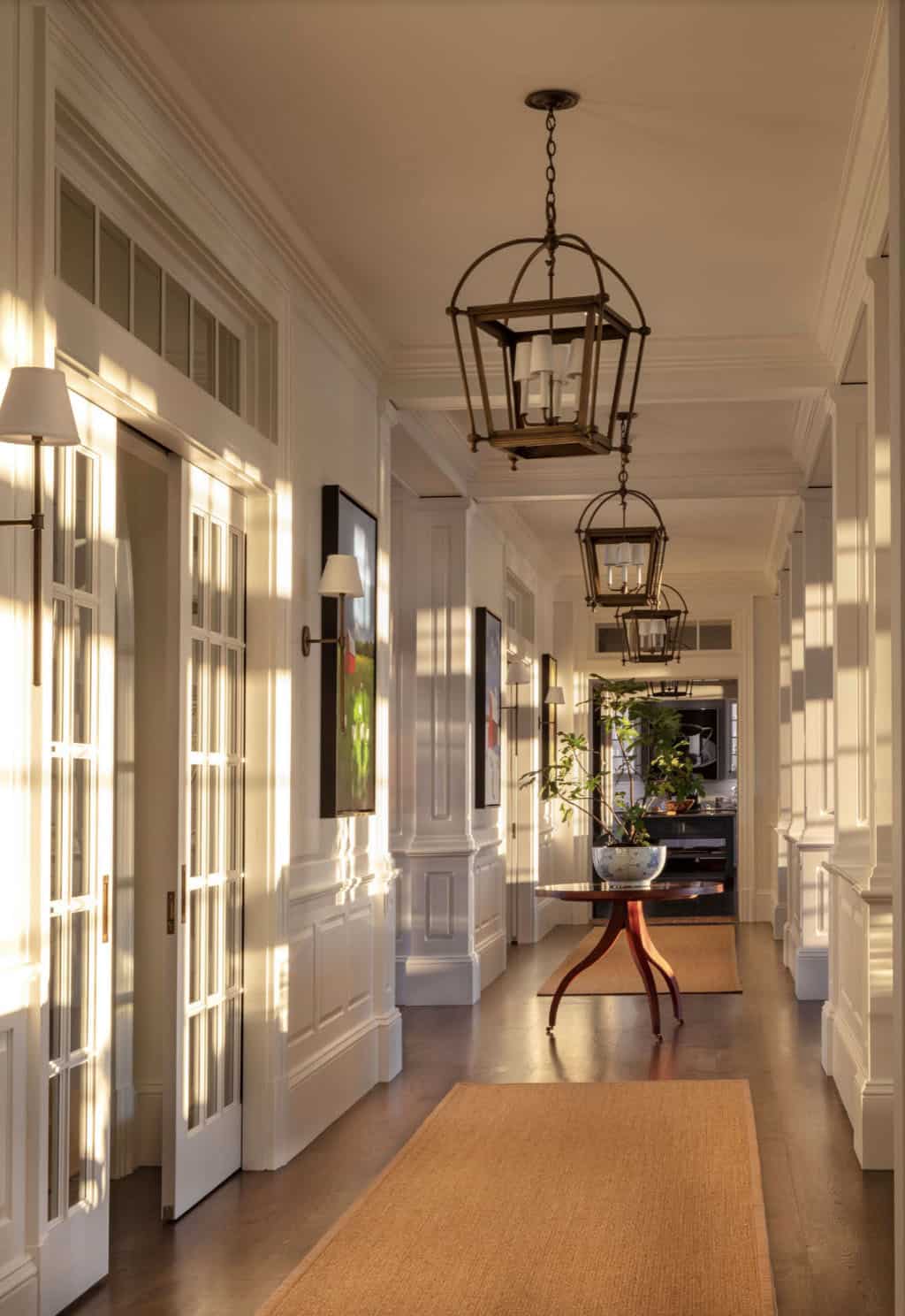 Sunlit interior hallway with repeating brass lantern pendants, wall sconces, paneled wainscoting, and a round pedestal table at the far end