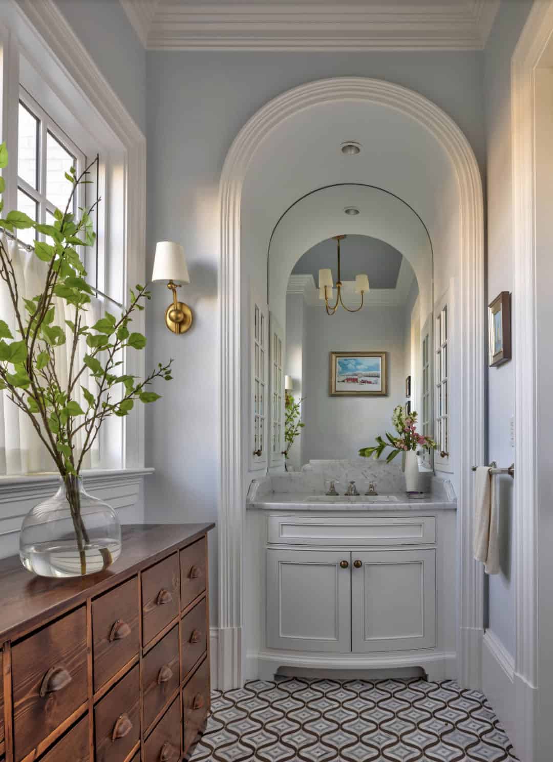 Powder room with arched niche vanity, patterned floor tile, marble countertop, brass sconce, and antique wood chest of drawers