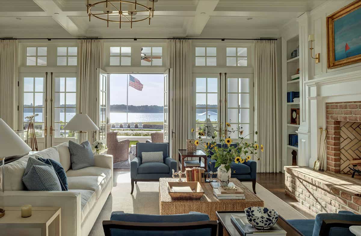 Waterfront sitting room with coffered ceiling, brass chandelier, white and blue furnishings, brick fireplace, and open French doors to the river