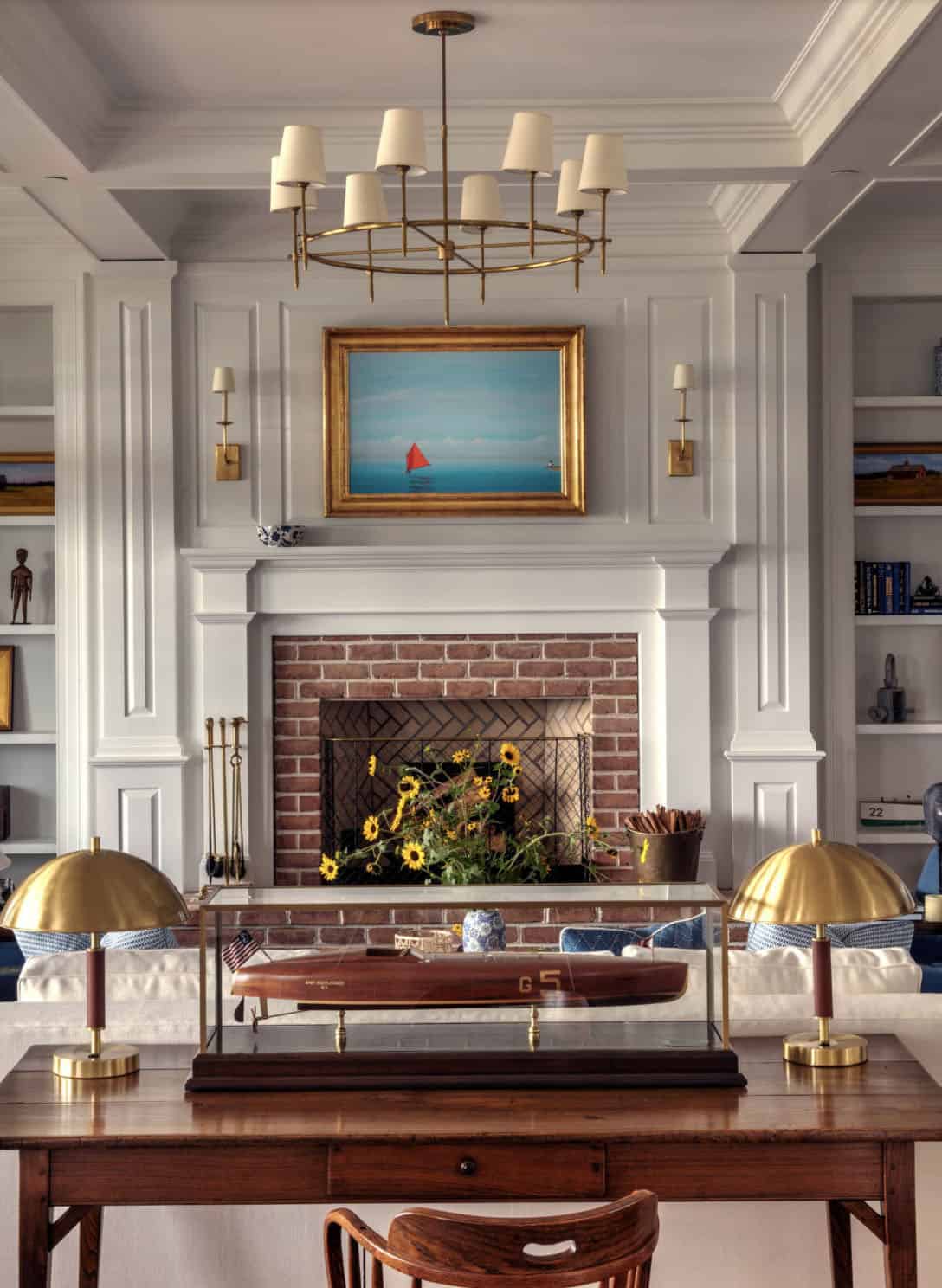Living room fireplace wall with white paneling, built-in bookshelves, brass ring chandelier, model boat display, and red sailboat painting above mantel