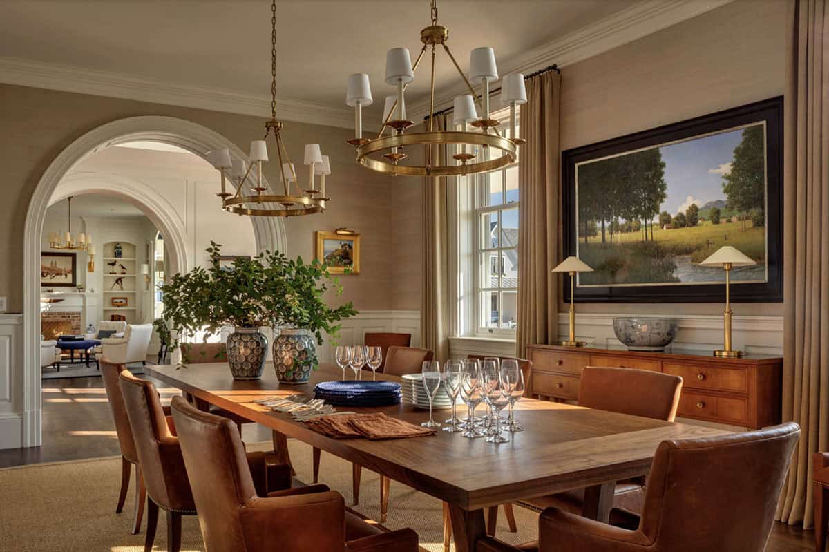 Formal dining room with walnut table, leather chairs, dual brass ring chandeliers, landscape painting, and arched passage to living room