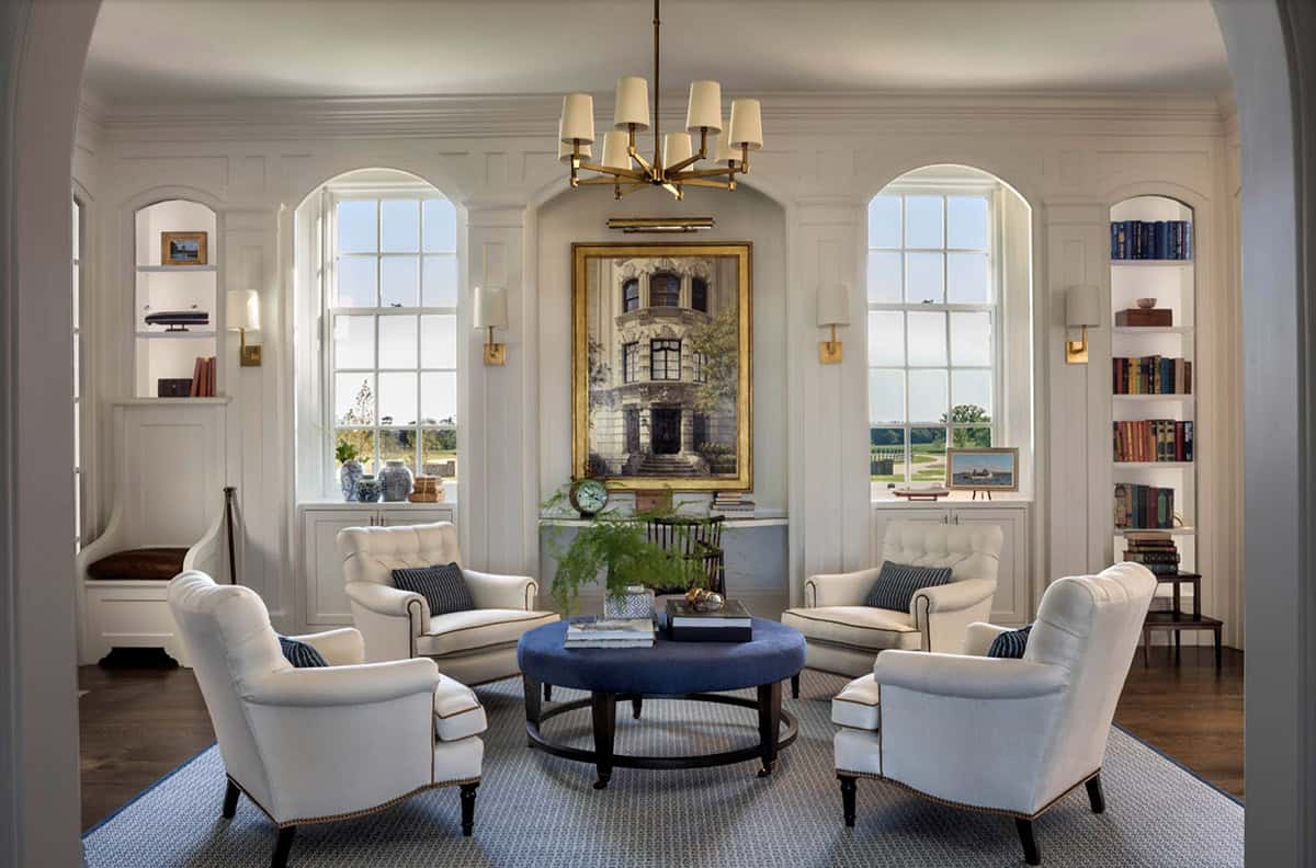 Elegant sitting room with four white tufted armchairs, navy ottoman, arched windows, brass chandelier, and large architectural painting