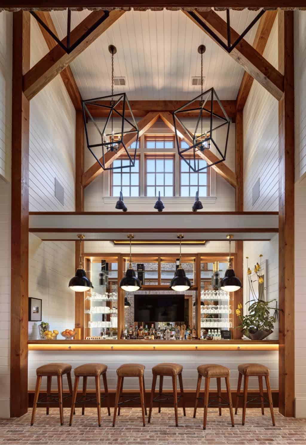 Dramatic barn bar with soaring timber-frame ceiling, geometric black pendant lights, backlit bar with leather stools, and brick floor