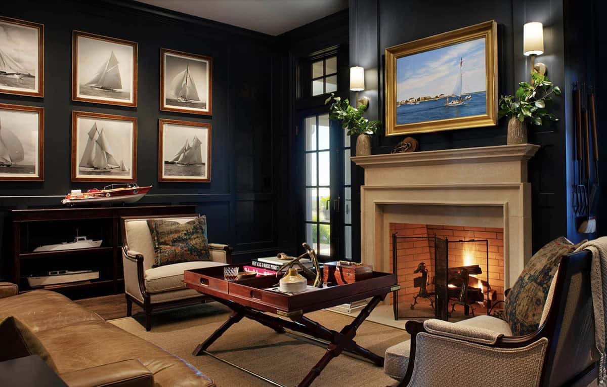Moody navy sitting room with lit fireplace, collection of black-and-white sailboat photographs, leather sofa, and nautical painting above mantel