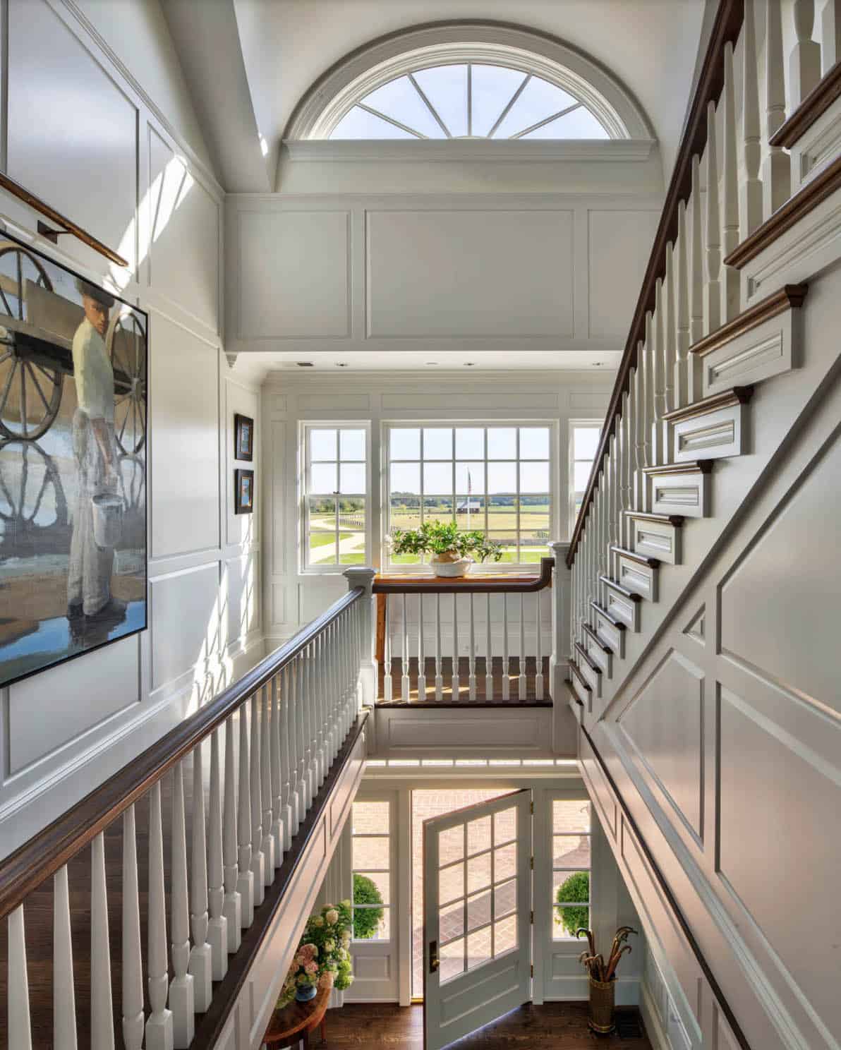 Upper stair hall looking down to front entry door with fanlight window, paneled walls, dark wood railings, and estate views beyond