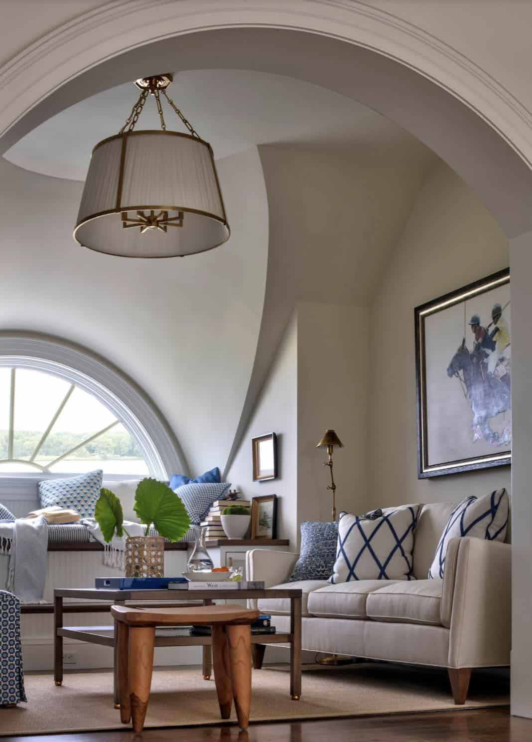 Cozy upper-floor sitting nook framed by an arch with fan window, linen sofa, blue pillows, and equestrian painting on the wall