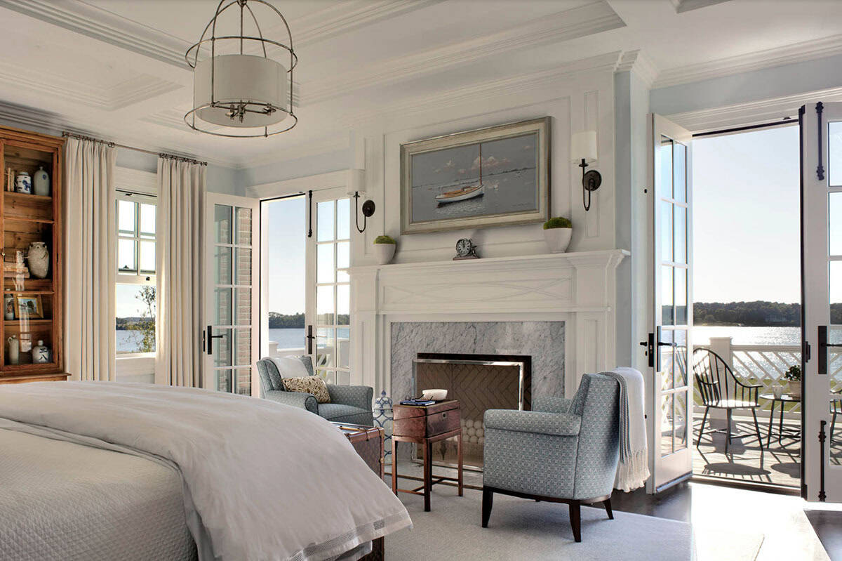Serene primary bedroom with marble fireplace, blue armchairs, sailboat painting, French doors open to waterfront balcony