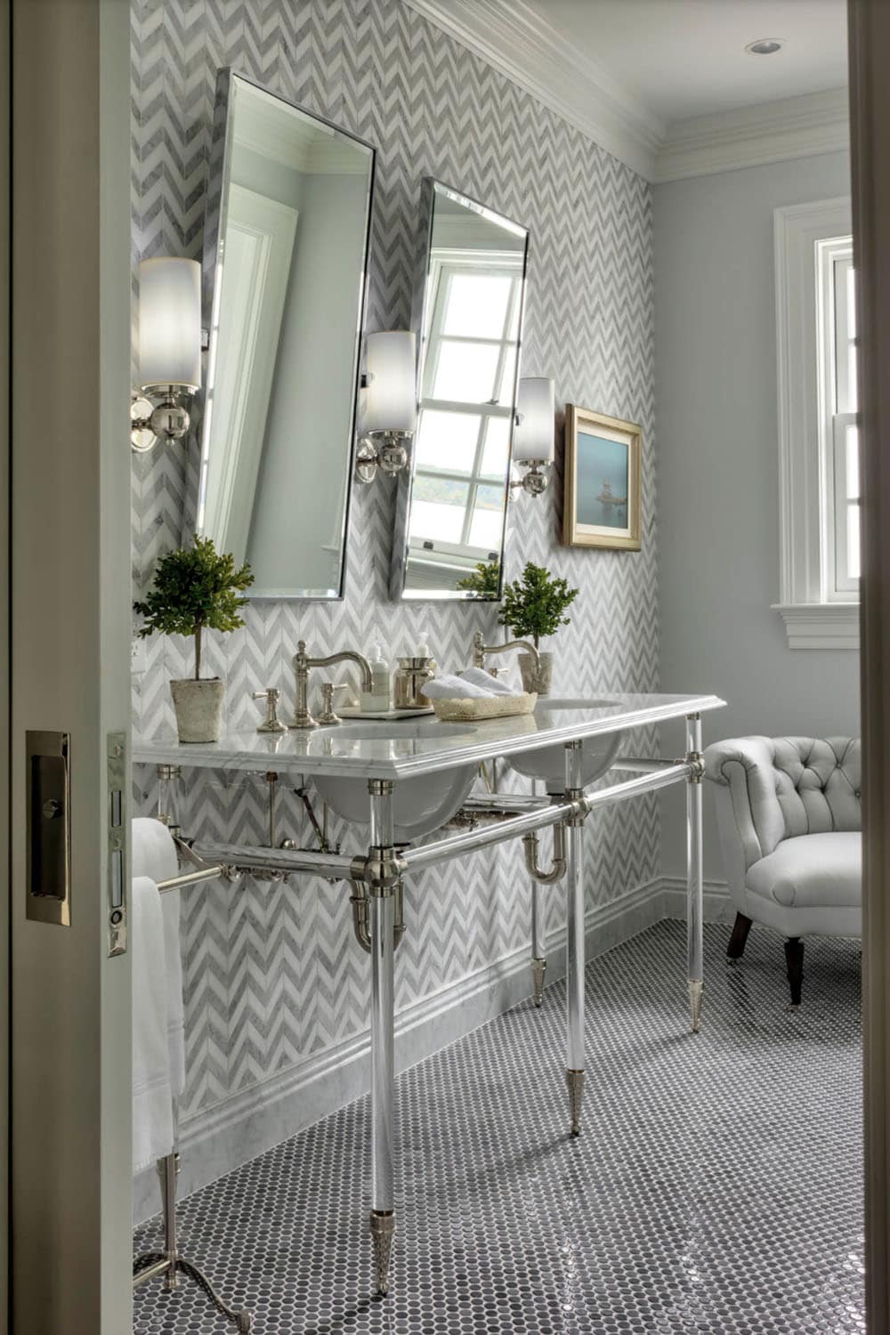 Primary bathroom with gray chevron marble tile walls, penny tile floor, polished nickel console sink, beveled mirrors, and tufted chair