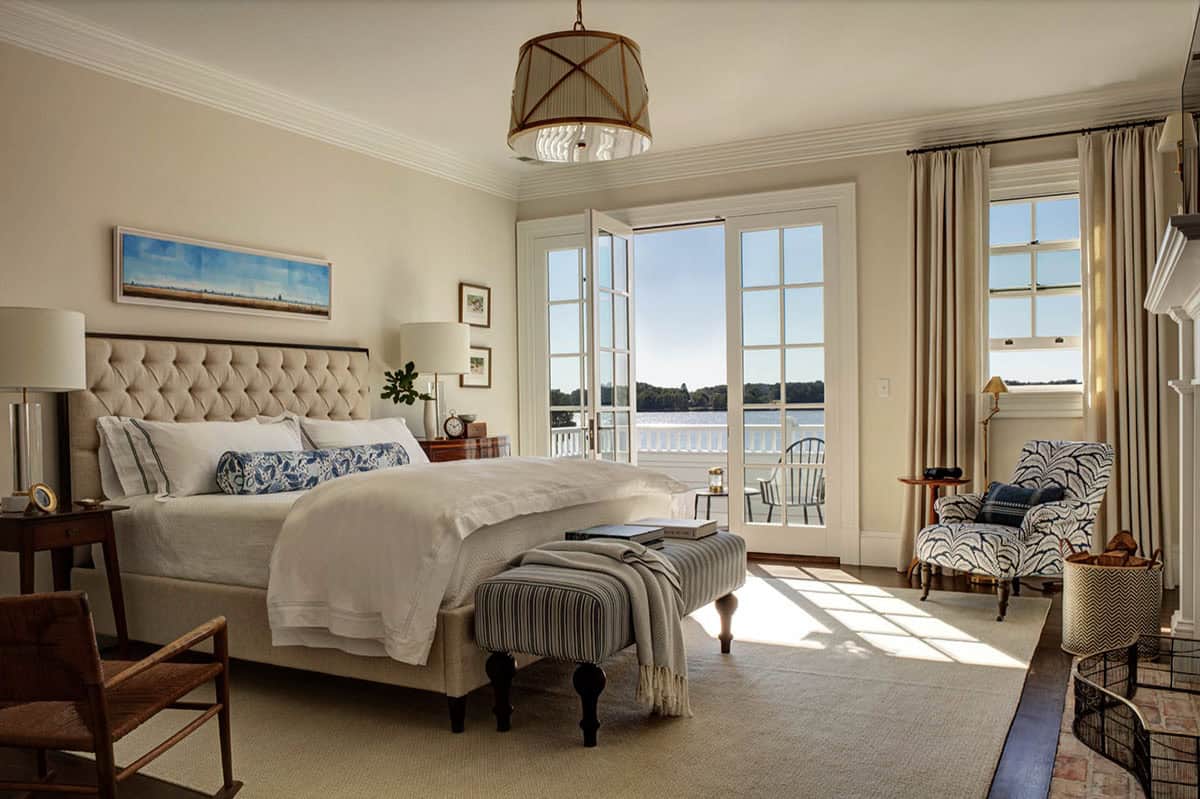Guest bedroom with tufted linen headboard, striped bench, French doors open to water view balcony, and blue botanical armchair