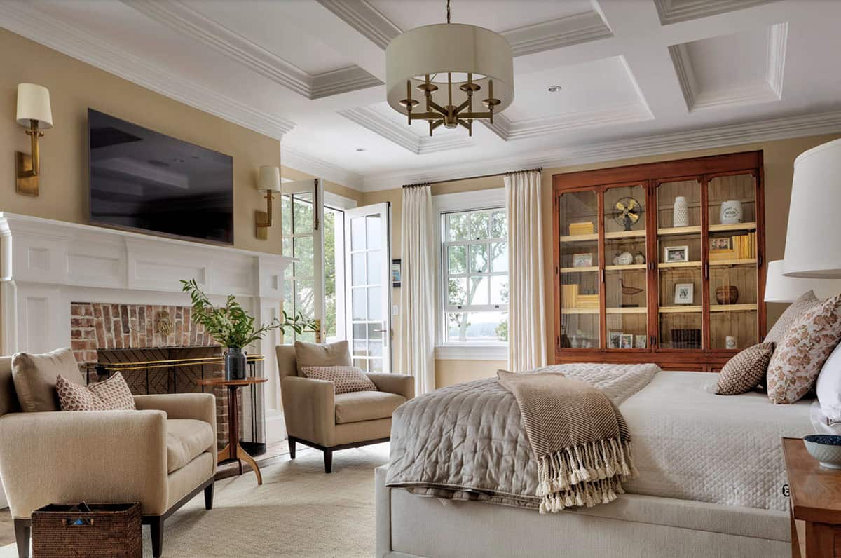 Warm guest bedroom with coffered ceiling, brick fireplace, antique glass-front cabinet, neutral upholstered furnishings, and waterfront windows